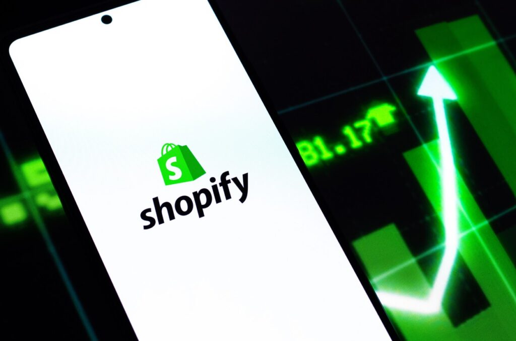 Shopify Logo auf einem Smartphone neben einem Aktienkurs